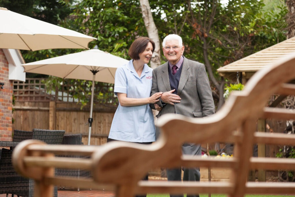 A B&M Bank Care worker walks arm in arm with an elderly gentleman in a care home garden.